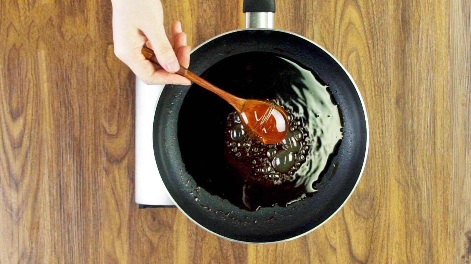 Tay cầm muỗng gỗ khuấy chất lỏng sẫm màu trong chảo trên mặt bàn gỗ.