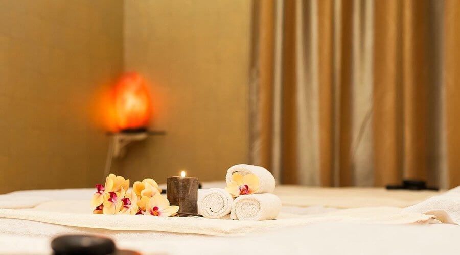 Khăn tắm cuộn và hoa lan trên giường massage, nến cháy sáng, nền là rèm cửa màu nâu, tạo không gian thư giãn.