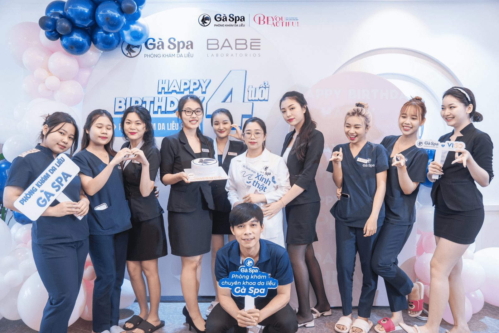 Một nhóm người chụp ảnh mừng sinh nhật với bánh kem và bóng bay, có bảng hiệu "Gà Spa".