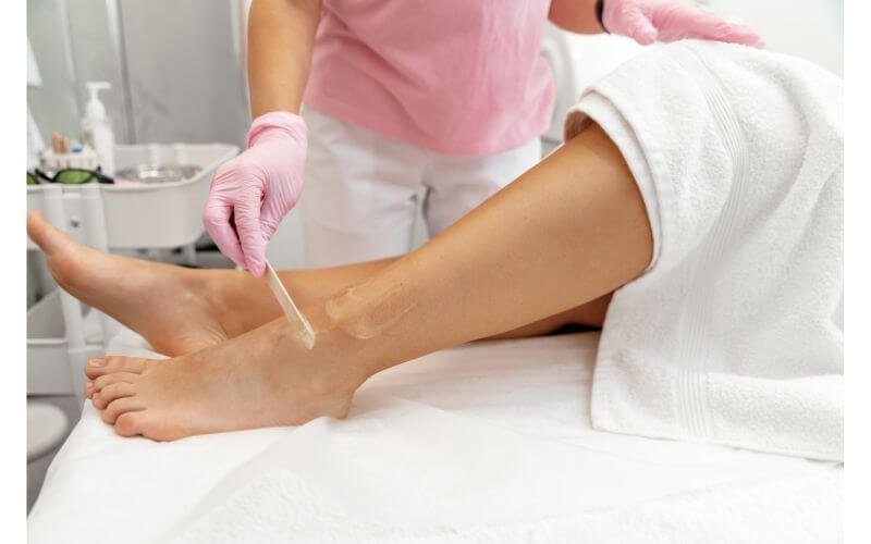 Người đang thoa kem trên chân của một khách hàng trong spa, tay đeo găng tay hồng, khăn trắng phủ chân.