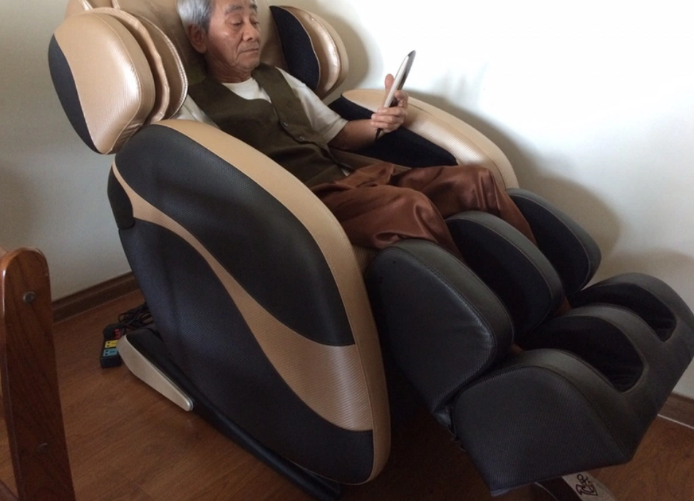 Người ngồi trên ghế massage, tay cầm điện thoại, trong phòng có sàn gỗ.