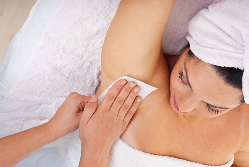 Người phụ nữ đang được tẩy lông nách bằng sáp, đầu quấn khăn, nằm trên khăn trắng trong spa.