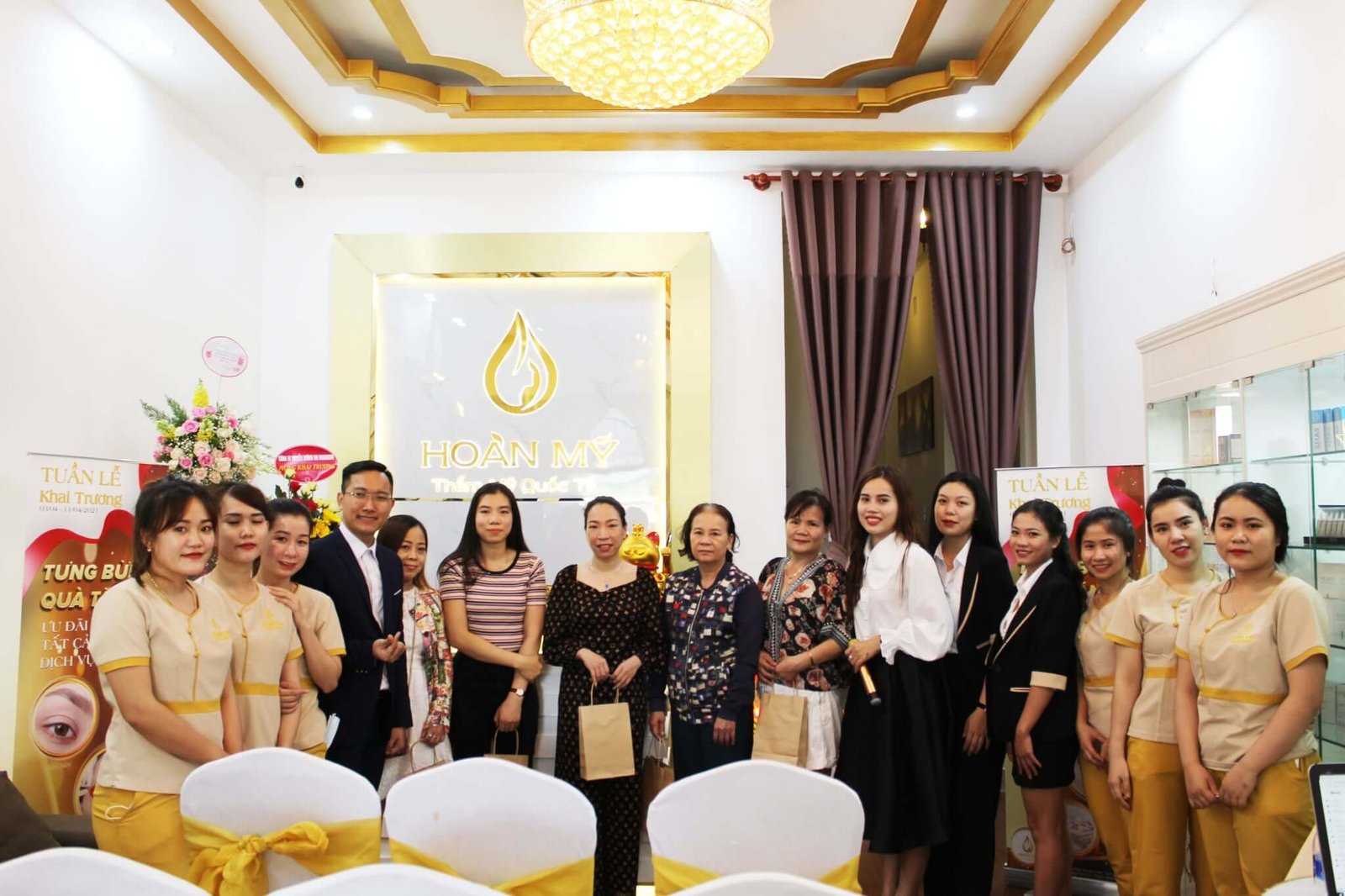 Nhóm người đứng cạnh nhau trong một buổi khai trương tại một thẩm mỹ viện có trang trí hoa và bảng hiệu.