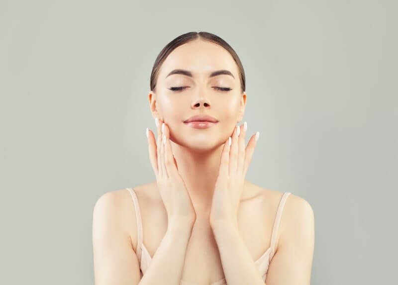 Người phụ nữ nhắm mắt, mỉm cười nhẹ, chạm tay lên mặt, nền xám trơn, không gian yên bình.