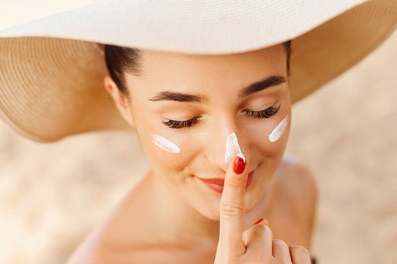 Người đội mũ rộng vành thoa kem chống nắng lên mũi, với nền cát mờ phía sau.