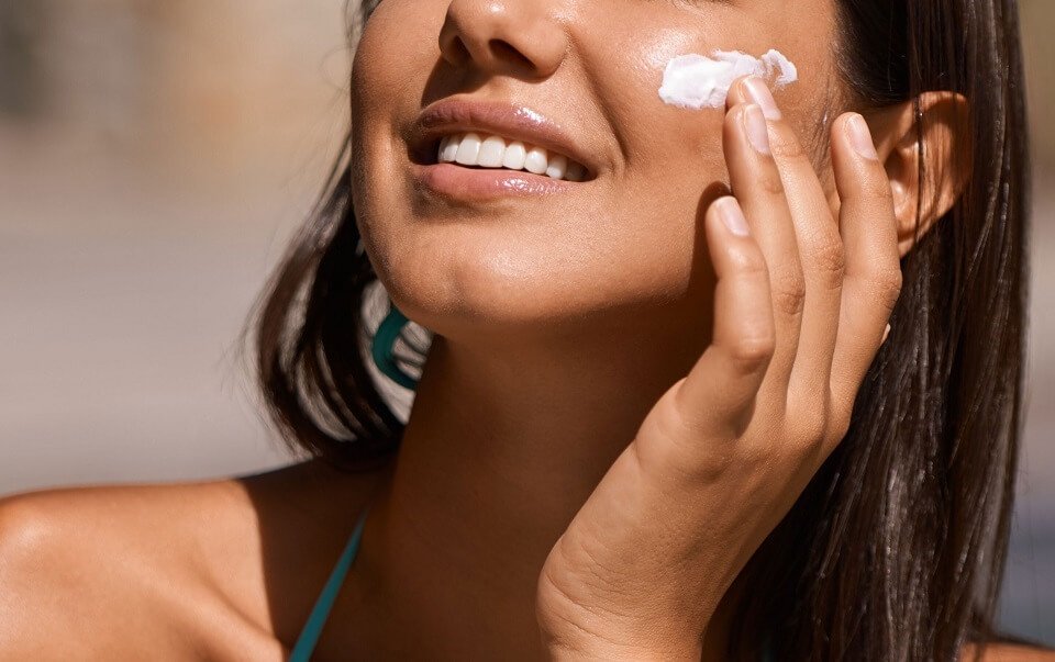 Người phụ nữ thoa kem dưỡng trên má, cười tươi, ánh sáng tự nhiên chiếu vào gương mặt.
