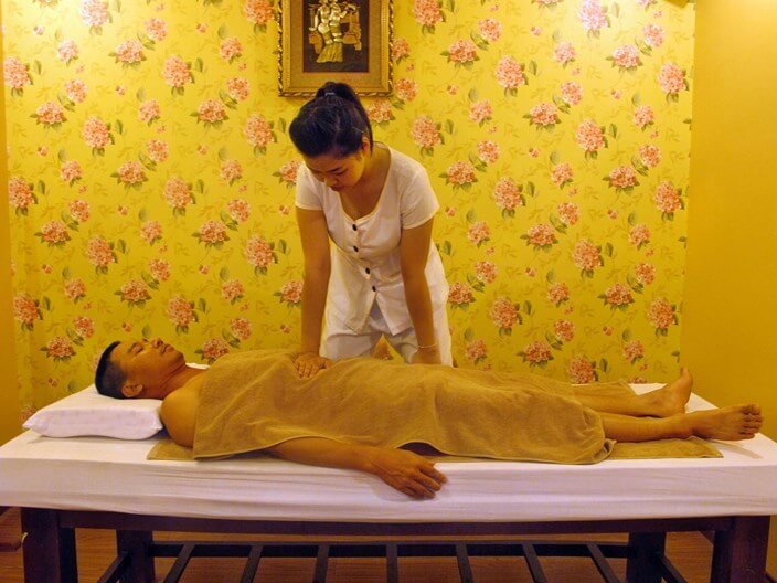 Người đang thực hiện massage cho một người nằm trên giường, trong phòng có giấy dán tường hoa.
