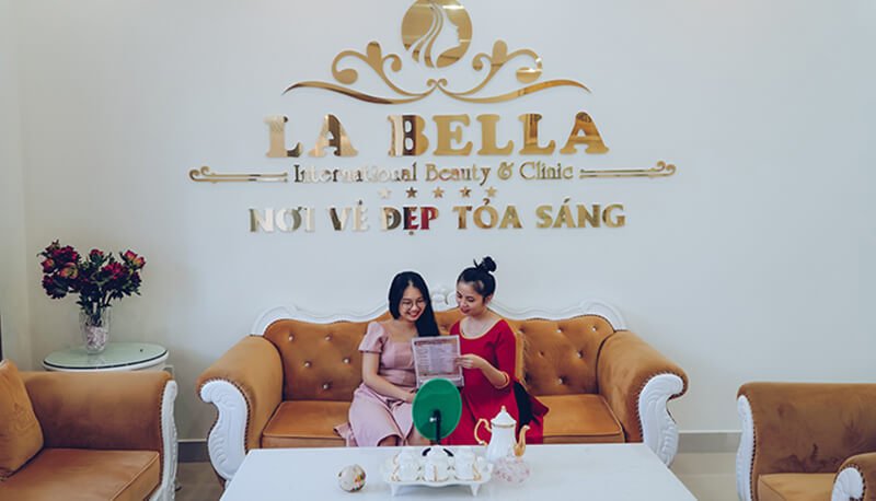 Hai người ngồi trên ghế sofa trong một thẩm mỹ viện, cùng xem tài liệu. Bố trí nội thất ấm cúng và sang trọng.