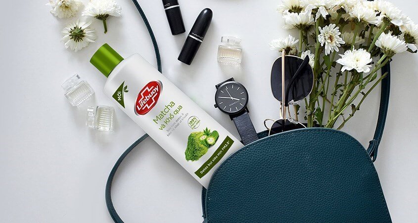 Chai sữa tắm matcha đặt cạnh túi xách xanh, hoa trắng, đồng hồ, kính mát và son môi trên nền trắng.
