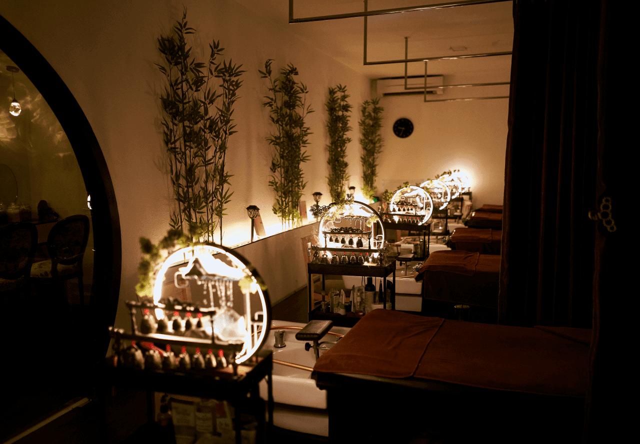 Phòng spa với giường massage, ánh sáng ấm áp, trang trí cây xanh và kệ chứa dầu massage.