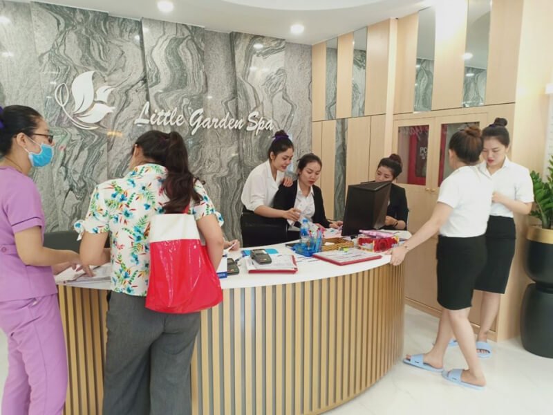Quầy lễ tân spa với nhân viên đang làm việc, khách hàng trao đổi và nội thất hiện đại, trang nhã.