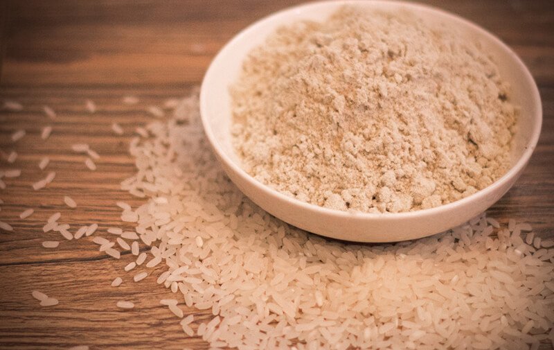 Bát bột gạo trên bàn gỗ, xung quanh có nhiều hạt gạo trắng rải rác.
