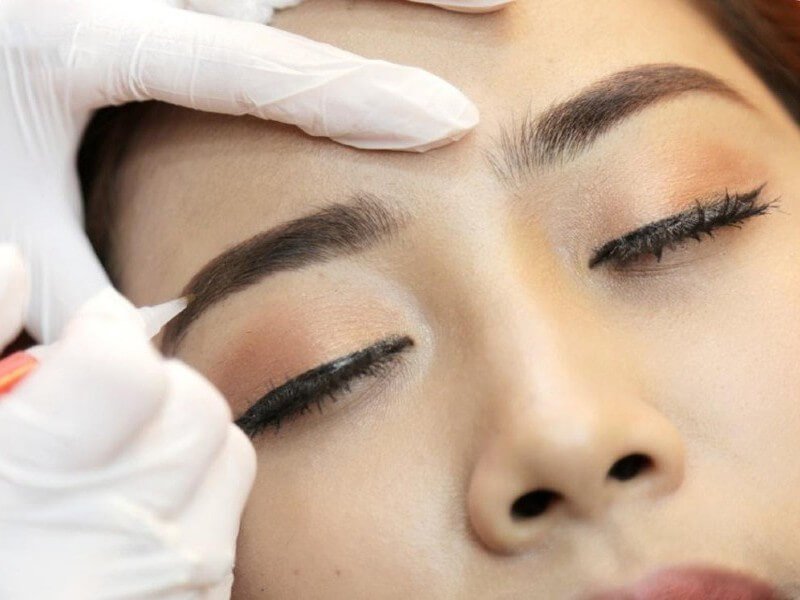 Người đeo găng tay trắng đang chỉnh sửa lông mày cho một người nhắm mắt.