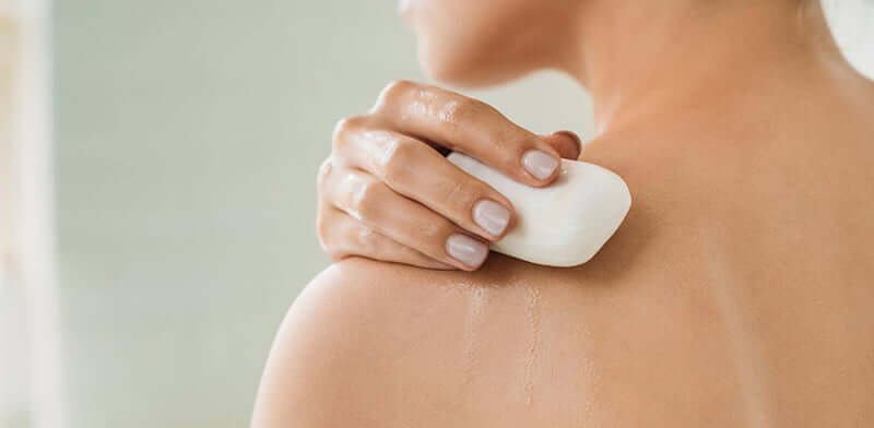 Người đang dùng xà phòng cục để tắm, xoa lên vai với nước chảy nhẹ trên da.