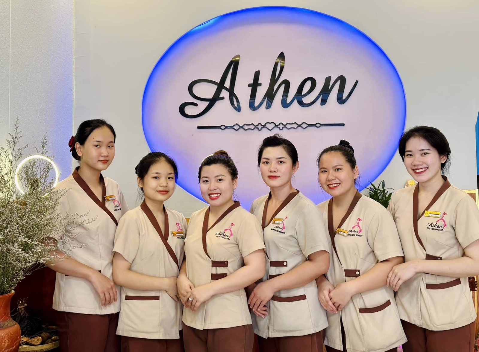 Nhóm người mặc đồng phục đứng trước bảng hiệu lớn trong không gian nội thất trang trí cây xanh.