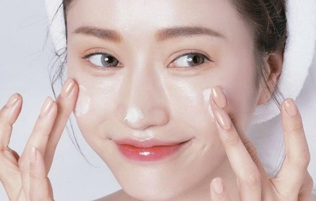 Người đang thoa kem dưỡng da lên mặt, với khăn quấn quanh đầu, trong bối cảnh sáng sủa.