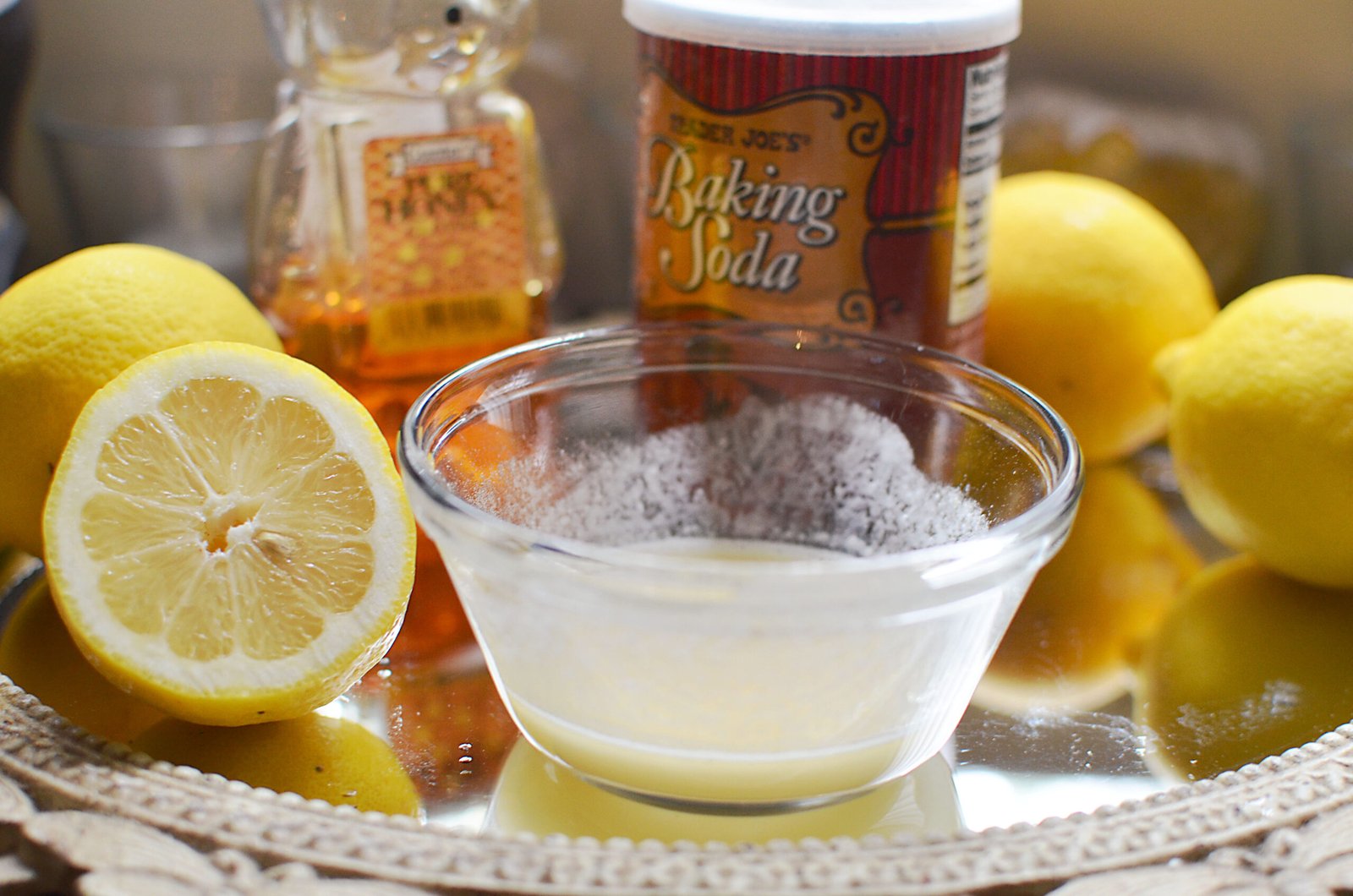 Chanh, mật ong, và baking soda trên bàn, với chanh cắt đôi và bát trộn gần đó.