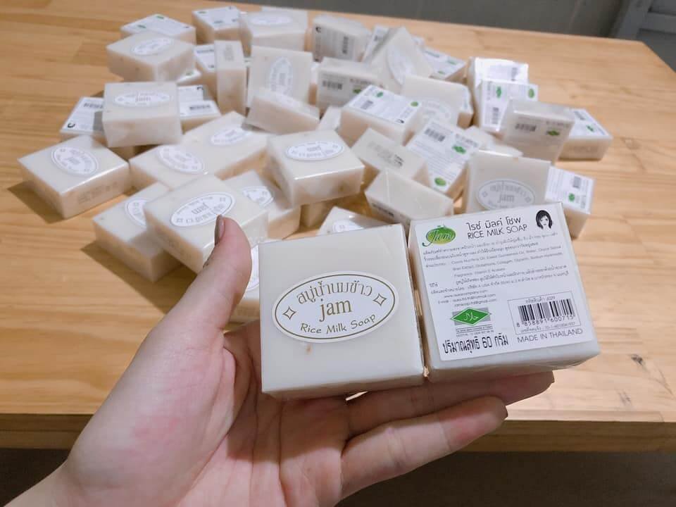 Người cầm hai bánh xà phòng sữa gạo trên tay, nhiều bánh xà phòng khác được xếp trên bàn gỗ.