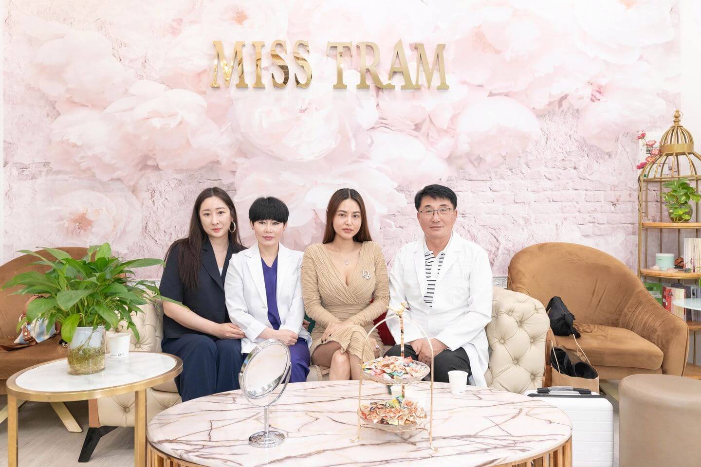 Bốn người ngồi trên ghế sofa trong phòng khách, nền hoa và chữ "MISS TRAM", có bàn và cây trang trí.