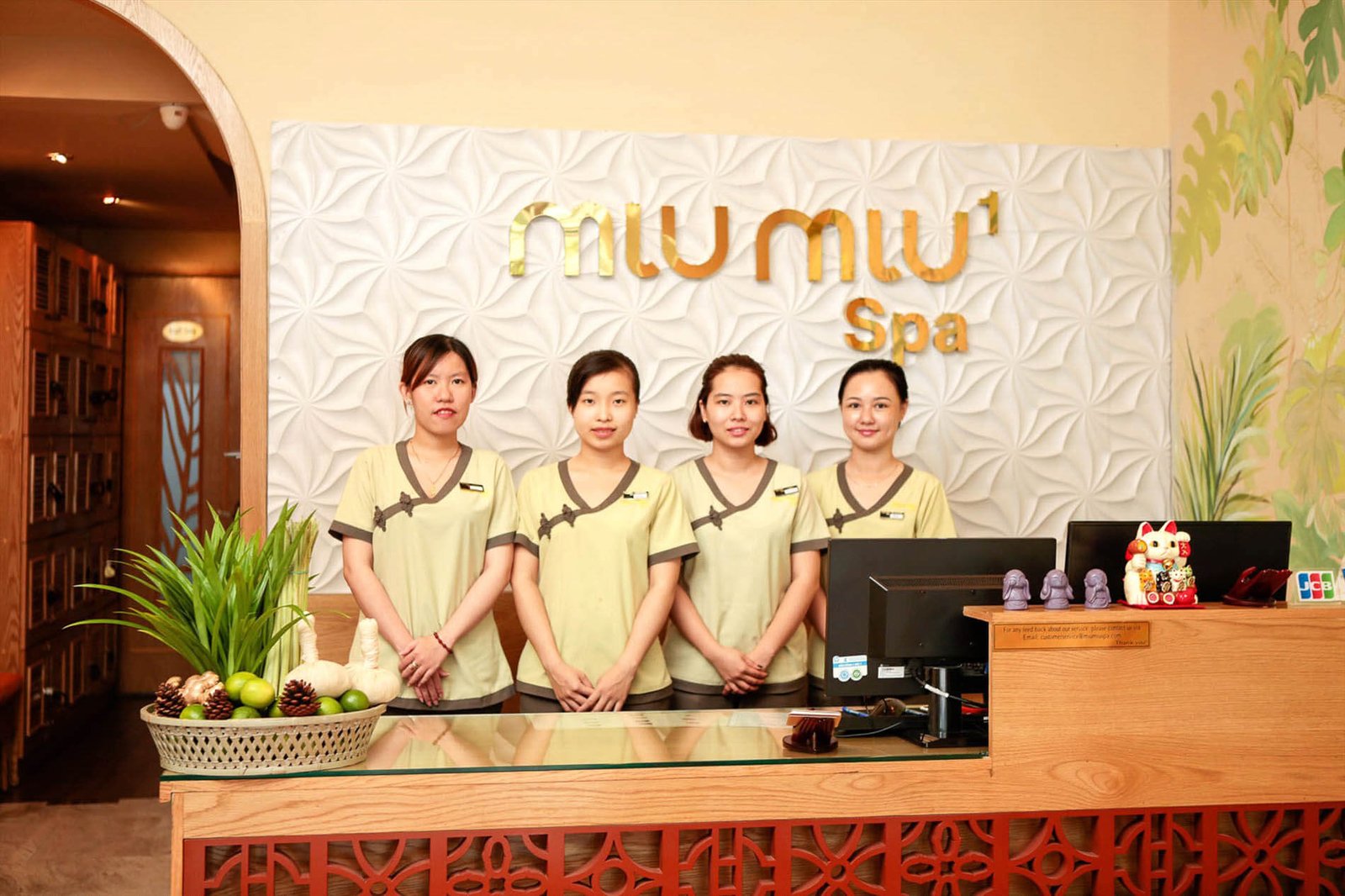 Bốn nhân viên spa đứng sau quầy lễ tân, phía sau là logo spa trên nền tường hoa văn.