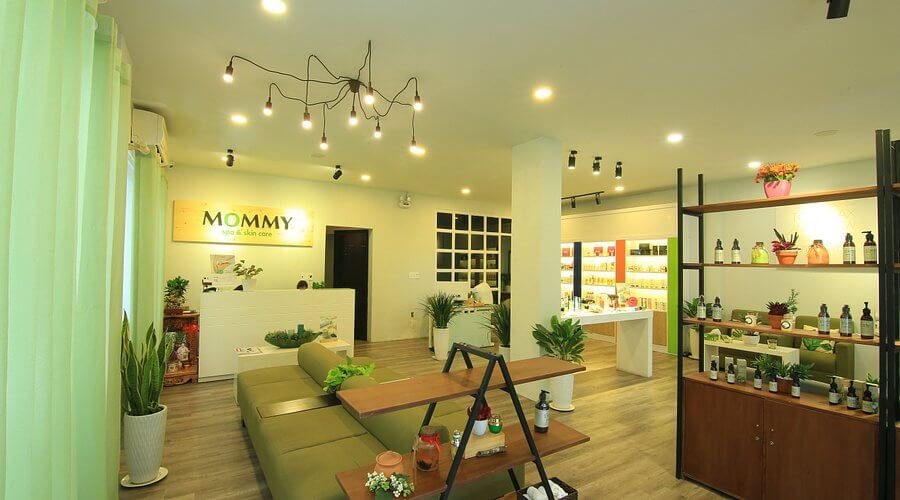 Phòng trang trí nội thất hiện đại với ghế sofa xanh, bàn gỗ, cây cảnh và kệ trưng bày sản phẩm dưới ánh sáng đèn.