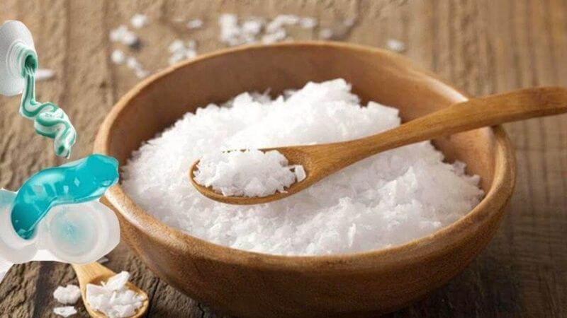 Bát gỗ chứa muối trắng, có thìa gỗ đặt bên trong. Bên cạnh là tuýp kem đánh răng màu xanh dương.