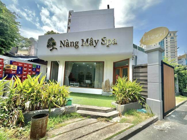 Mặt tiền một spa với bảng hiệu lớn, cây xanh trang trí trước cửa, nền trời xanh và nhiều mây.
