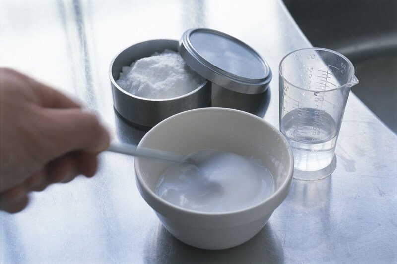 Người đang khuấy hỗn hợp trong bát, bên cạnh là hộp bột và cốc nước trên bàn kim loại.