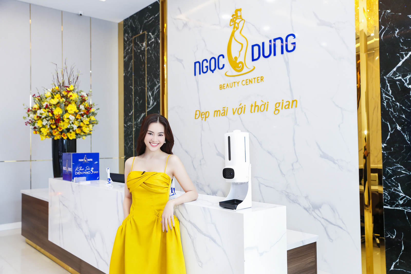 Người phụ nữ mặc váy vàng đứng cạnh quầy lễ tân trong trung tâm thẩm mỹ hiện đại với hoa trang trí.
