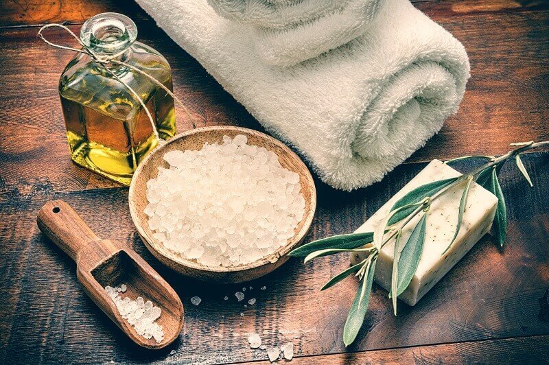 Tấm gỗ với khăn tắm trắng, chai dầu, muối tắm, xà phòng và nhánh cây ôliu.
