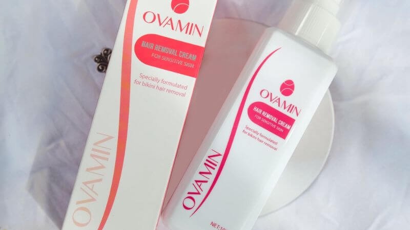 Hộp và chai kem tẩy lông nhãn hiệu Ovamin, nền trắng, chữ và họa tiết màu đỏ.