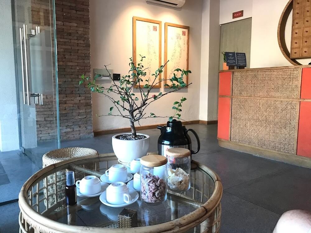 Bàn tròn với ấm trà, chén tách và cây cảnh trong không gian nội thất ấm cúng, có quầy lễ tân ở phía xa.