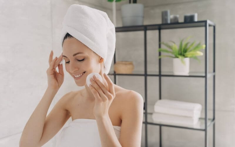 Người phụ nữ quấn khăn trên đầu, mỉm cười và dùng bông tẩy trang, đứng trong phòng tắm hiện đại với kệ và cây xanh.