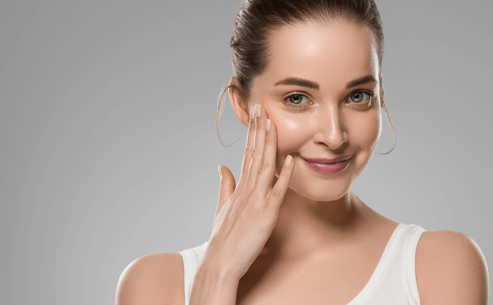 Người phụ nữ với làn da mịn màng, chạm nhẹ vào má, nền xám, mặc áo không tay màu trắng.