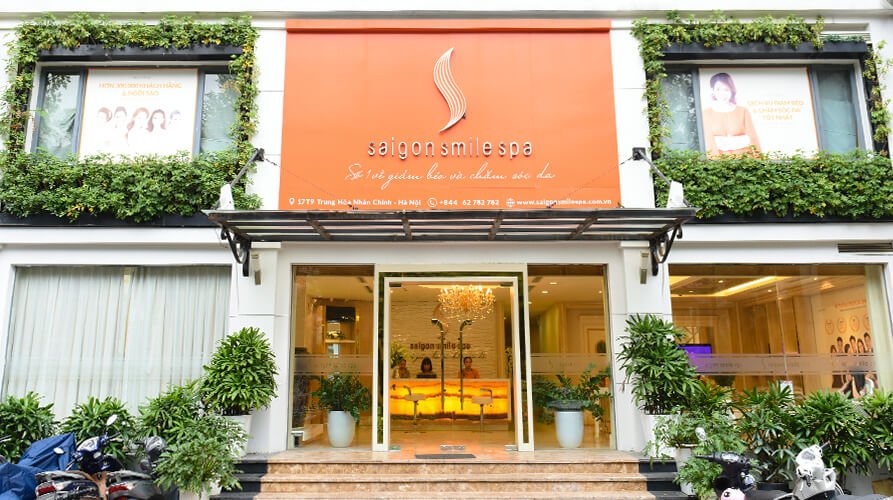 Mặt tiền một spa với cửa kính, bảng hiệu cam, cây xanh trang trí và xe máy đậu phía trước.