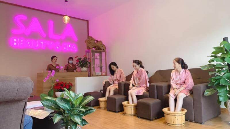 Ba người mặc áo choàng ngồi ngâm chân trong bồn gỗ tại một spa, phía trước quầy lễ tân có biển hiệu neon.
