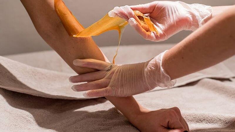 Quá trình tẩy lông bằng sáp trên cánh tay, người đeo găng tay đang thực hiện trên nền vải màu be.