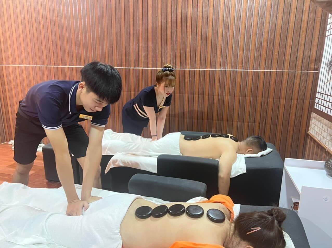 Hai người đang nhận liệu pháp massage đá nóng trong phòng trị liệu với hai nhân viên hỗ trợ.