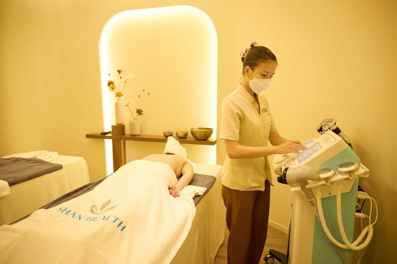 Nhân viên spa đang điều chỉnh máy móc, khách hàng nằm trên giường massage trong phòng ấm cúng với ánh sáng dịu.