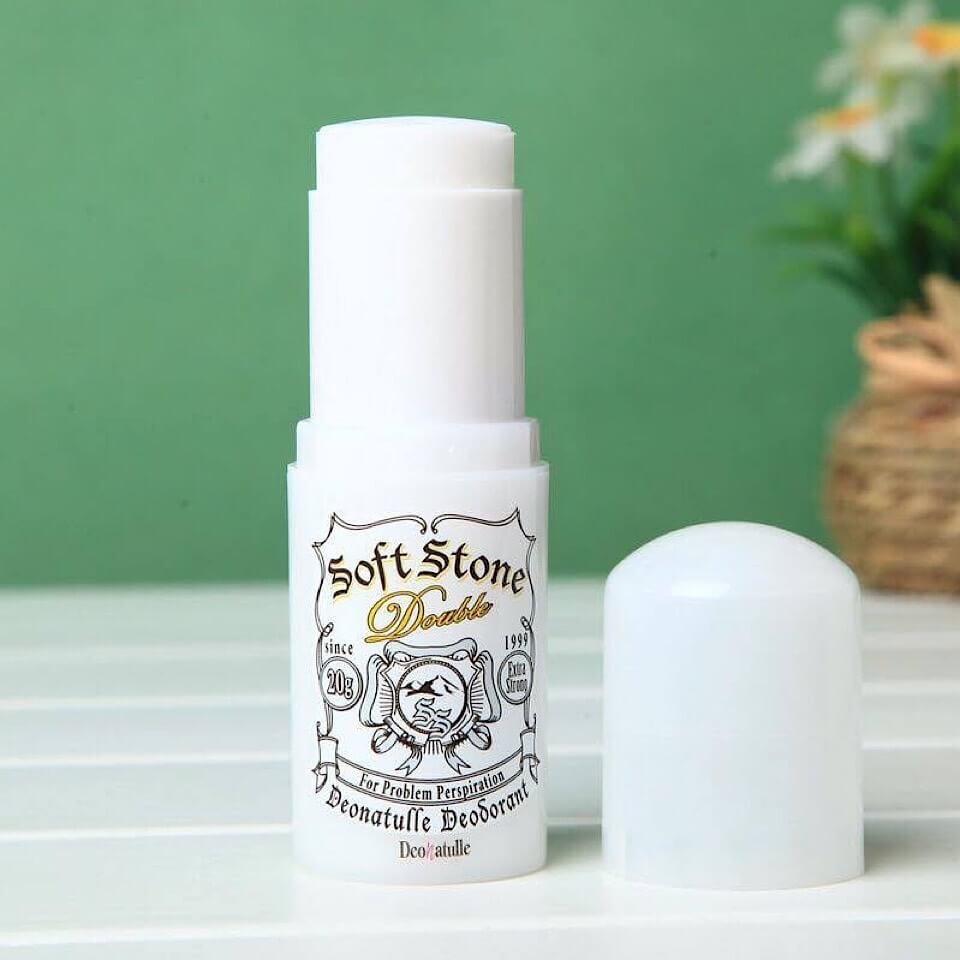 Lăn khử mùi dạng thỏi màu trắng đặt trên bàn, nắp mở, nền xanh lá cây và một giỏ hoa mờ phía sau.