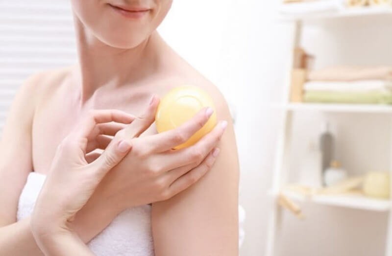 Người đang sử dụng quả bóng tròn màu vàng để massage vai trong phòng tắm, với kệ đựng đồ phía sau.