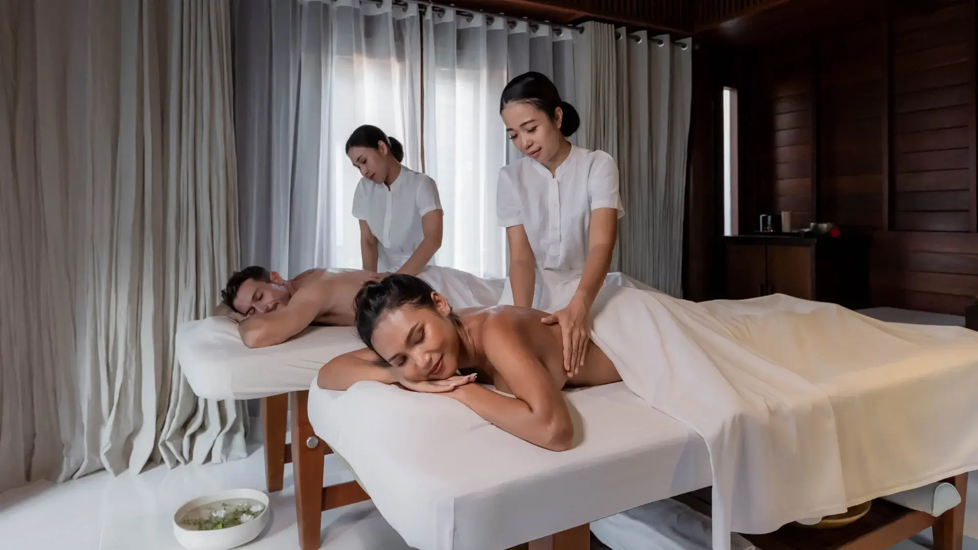 Hai người đang được mát-xa trên giường trong một phòng spa, có rèm trắng và ánh sáng dịu dàng.