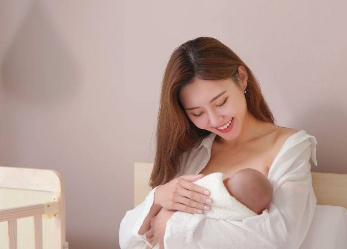 Người phụ nữ đang ôm một em bé sơ sinh bọc trong chăn, ngồi trên giường cạnh chiếc cũi gỗ.