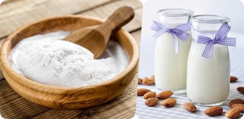 Bát bột trắng với muỗng gỗ bên trái, hai chai sữa kèm nơ tím và hạnh nhân trên bàn bên phải.