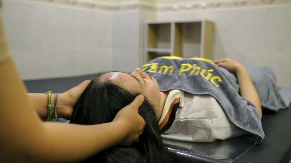 Người nằm thư giãn trên giường massage, có khăn phủ trên người, được mát-xa đầu trong không gian tĩnh lặng.