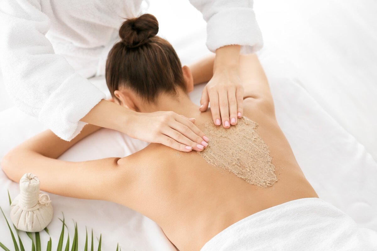 Người đang được mát-xa lưng bằng hỗn hợp tẩy tế bào chết trong spa, cạnh đó có túi thảo dược và lá cây.