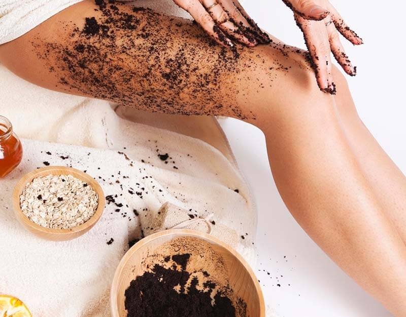 Người đang thoa hỗn hợp tẩy tế bào chết từ cà phê lên chân, bên cạnh có bát yến mạch và mật ong.