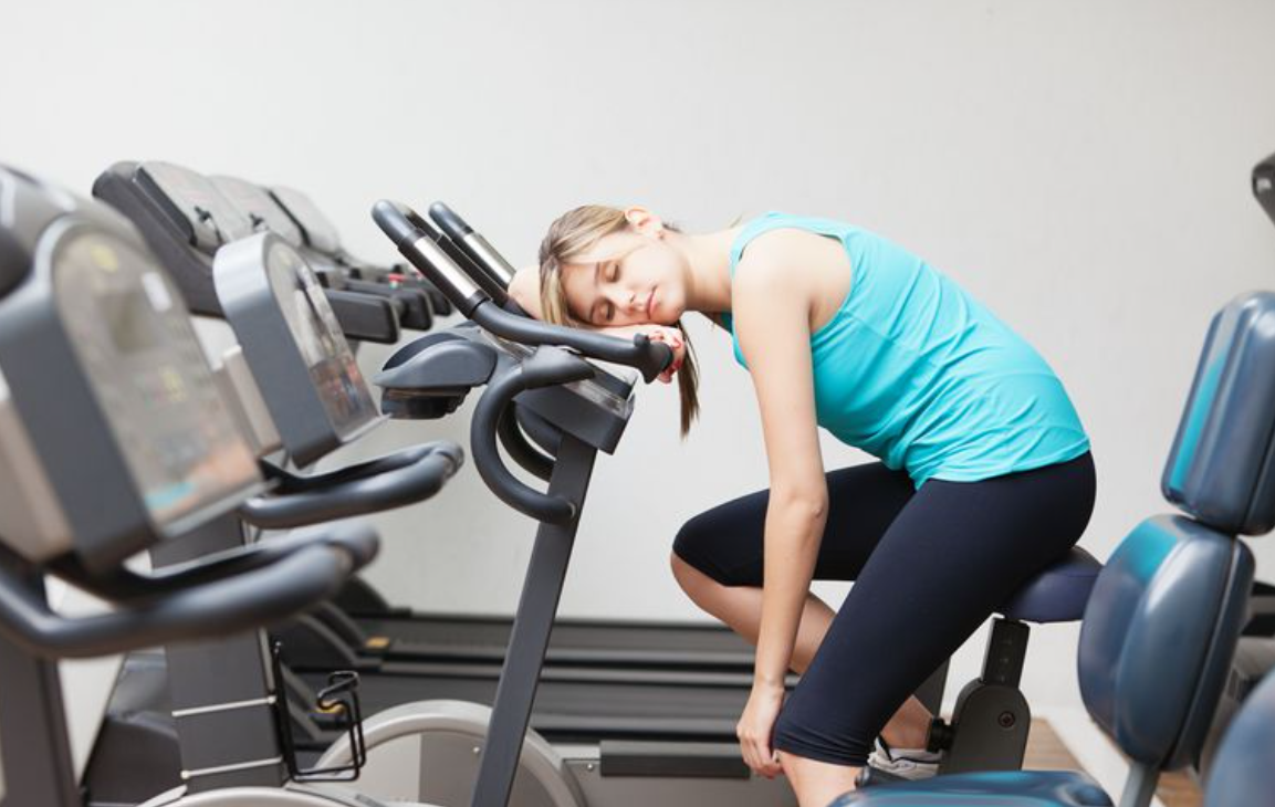 Người phụ nữ mặc đồ thể thao xanh dương, ngủ gục trên máy tập thể dục trong phòng gym.