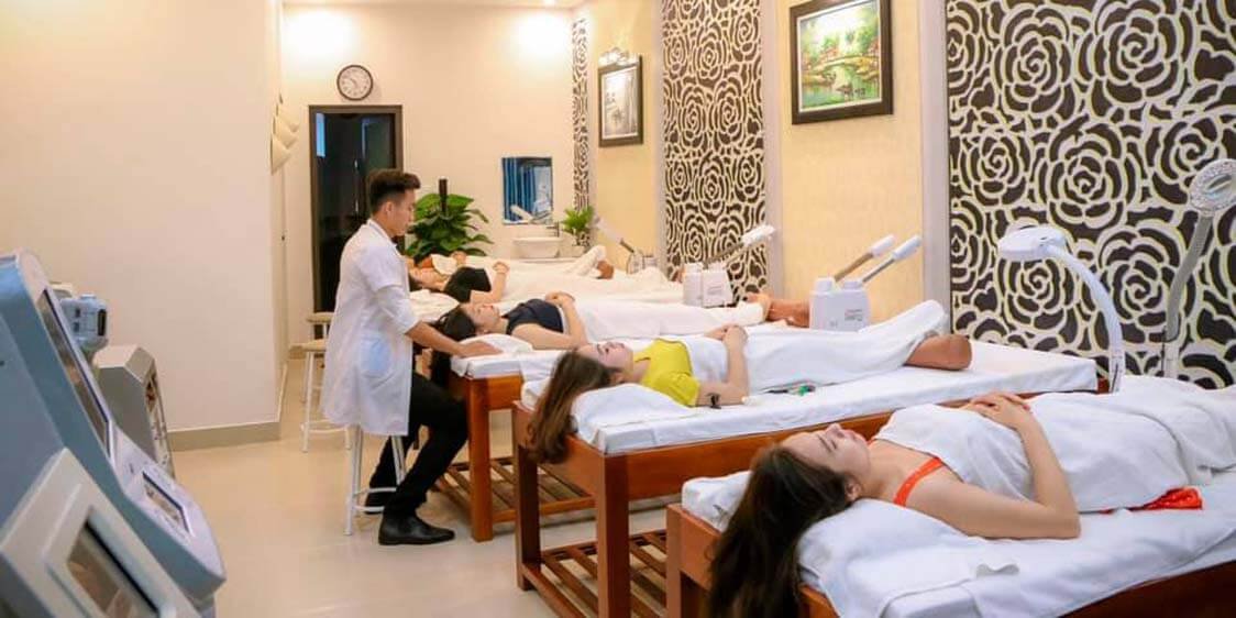 Phòng spa với nhiều giường massage, có người nằm thư giãn và một nhân viên đang chăm sóc khách hàng.