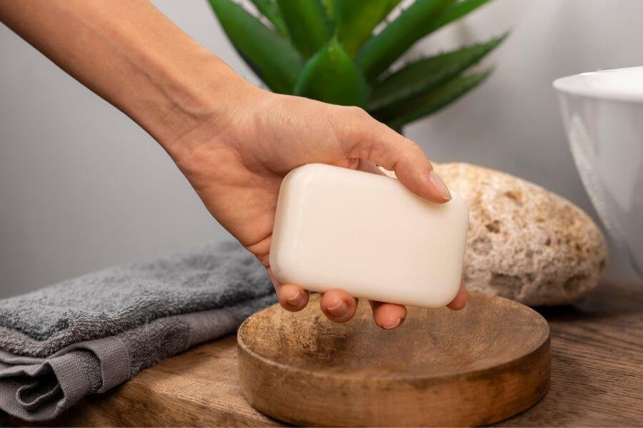 Tay cầm bánh xà phòng trắng trên đĩa gỗ, cạnh đó là khăn tắm và cây xanh.
